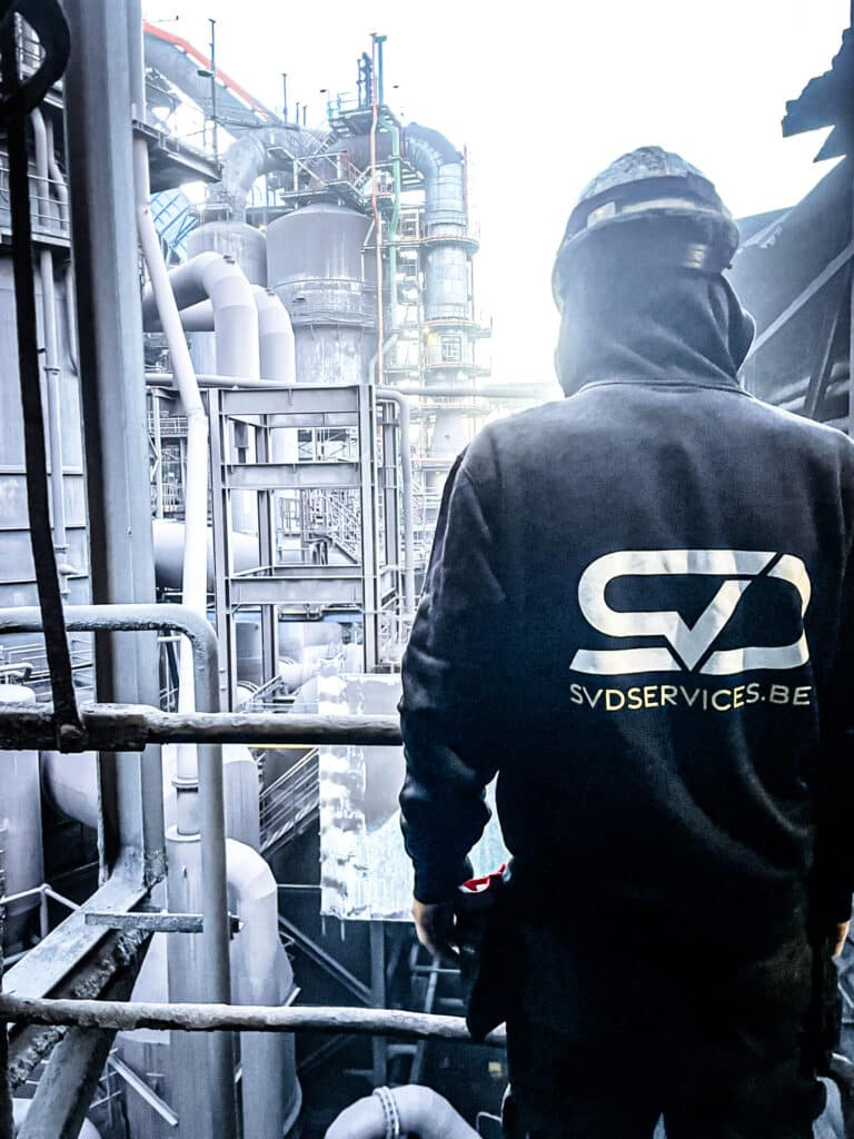 SVD Services monteur in branded hoodie inspecteert industriële installatie met buizen en torens in petrochemische omgeving
